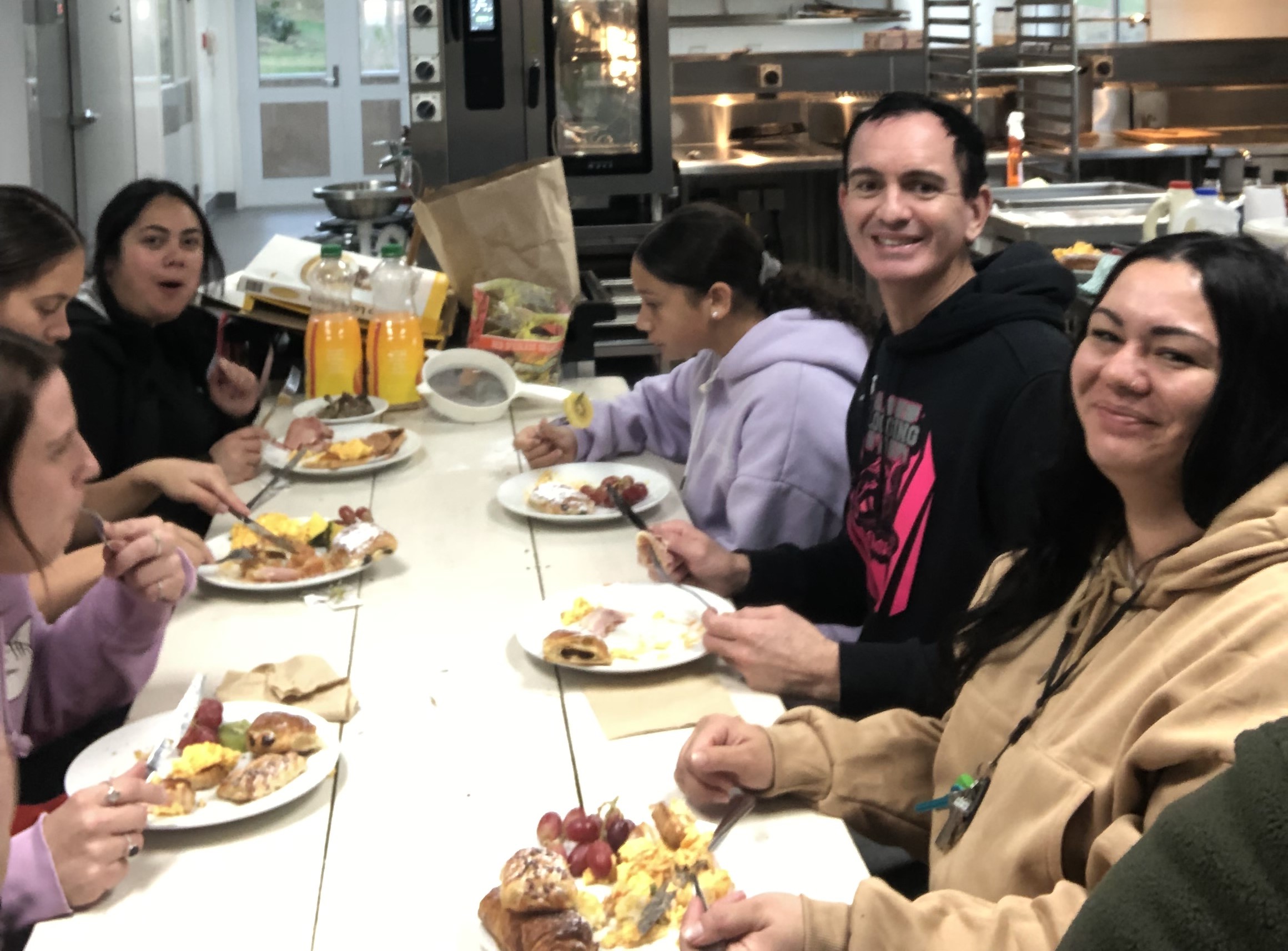 Several people at a table with full plates of breakfast foods eating and smiling at the camera.  