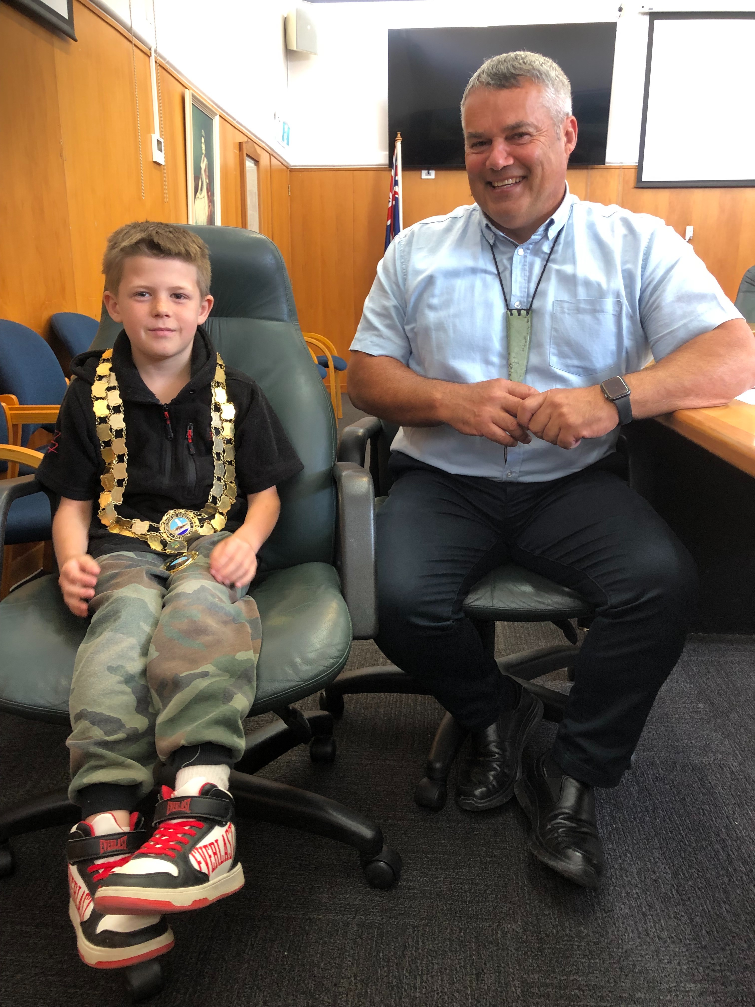 Young boy wearing mayoral chains sits in council chair with the mayor alongside. Both smiling. 