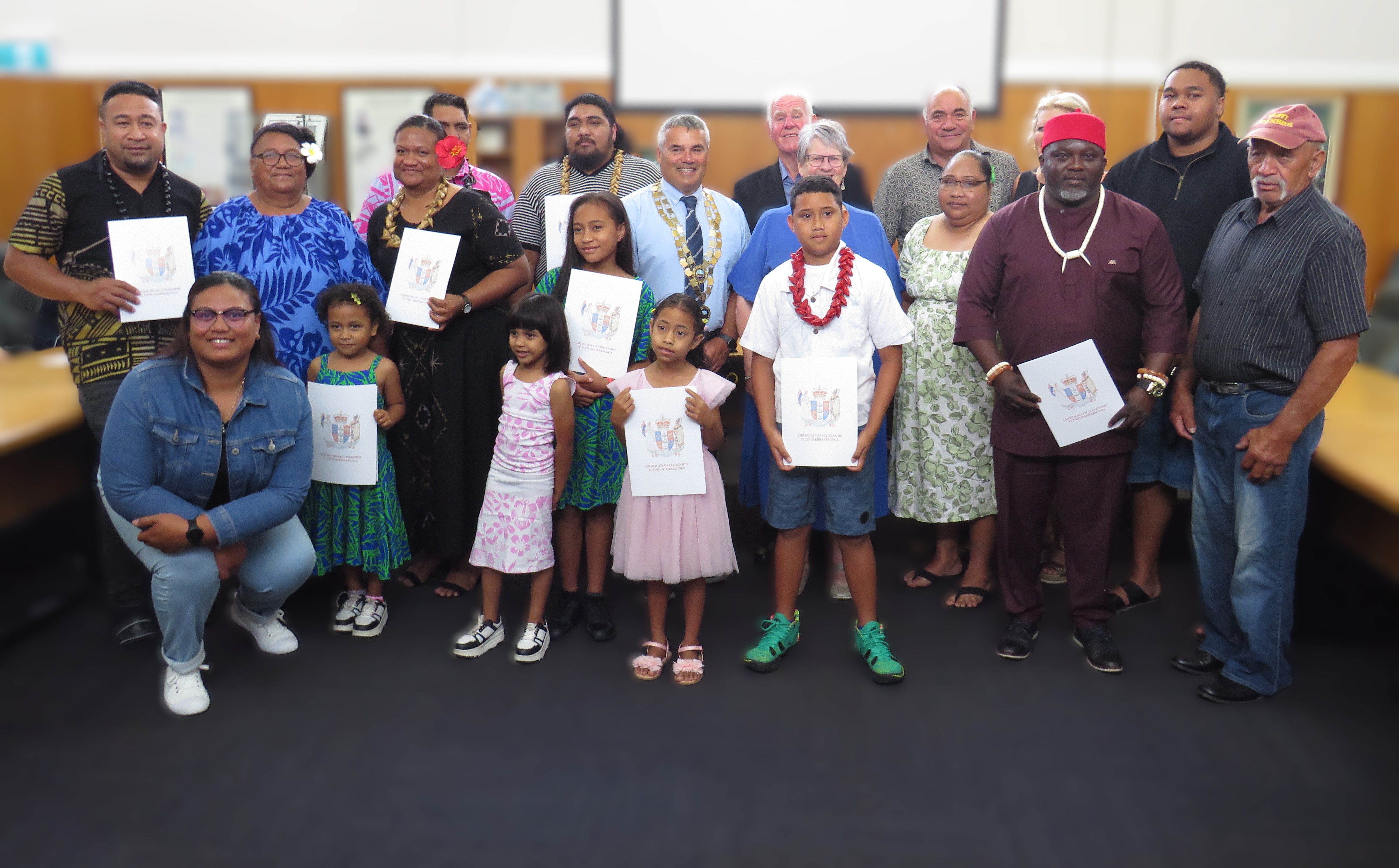 Group photo of twenty adults and children proudly holding their new citizenship certificates with the Mayor in his chains of office in the centre of the photo.