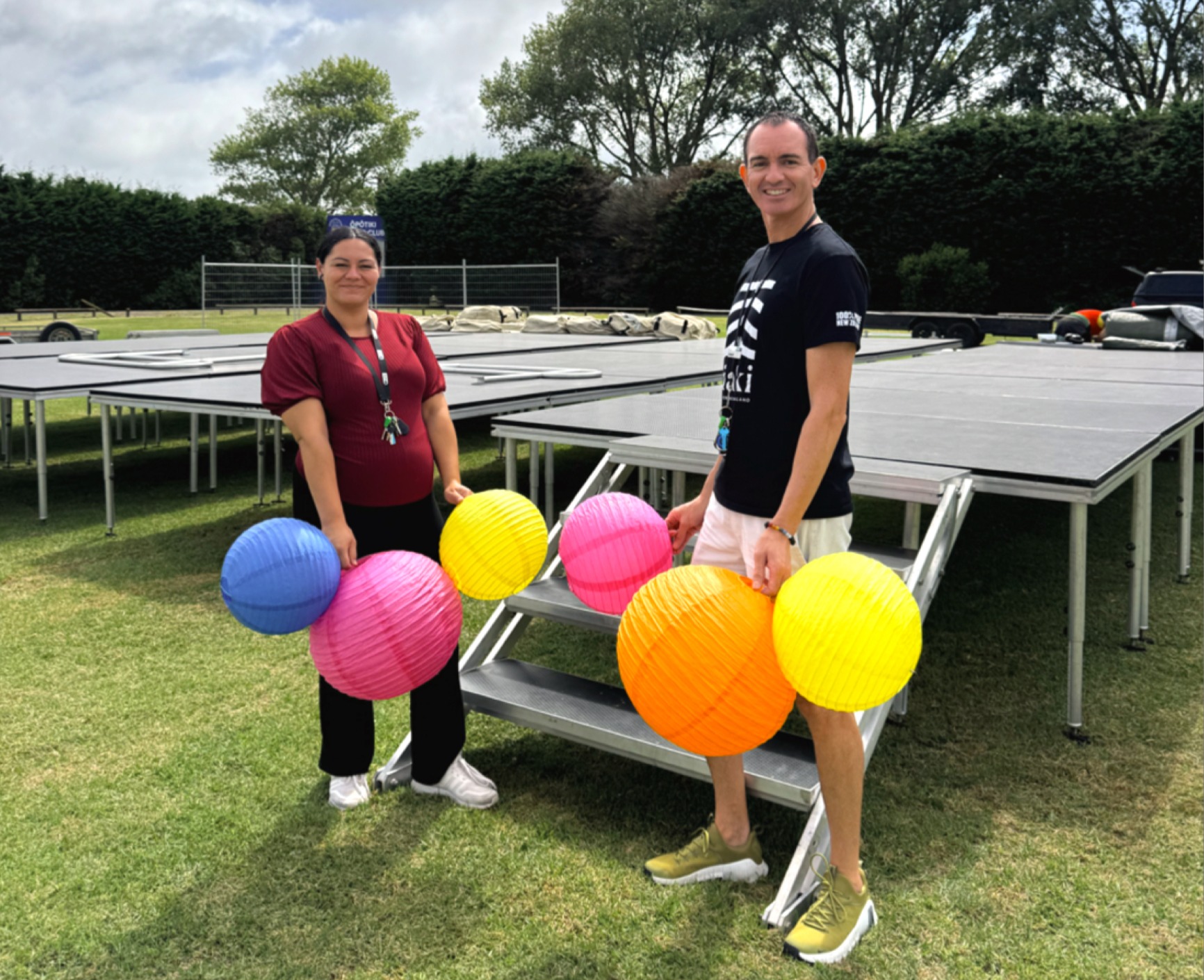 Young woman and young man stand in front of half-erected stage holding colourful lanterns. 