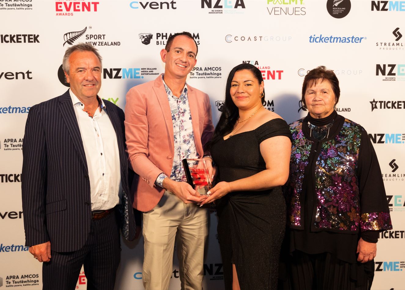 Event photo of four finely-dressed people holding an award trophy with lots of sponsorship logos on the wall behind. 