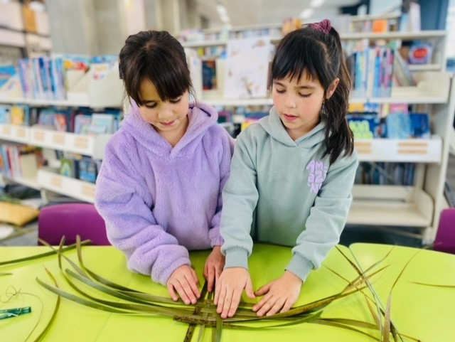 Two young girls work together to weave harakeke flax. 