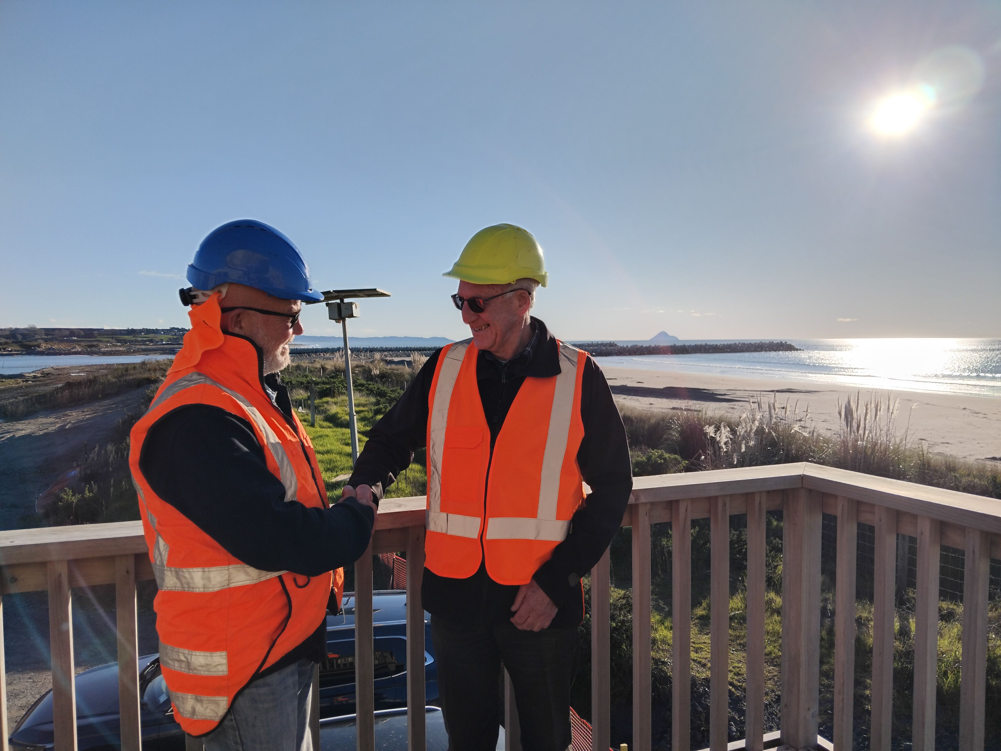 Two men in high viz shake hands at site office overlooking the Opotiki harbour seawalls.